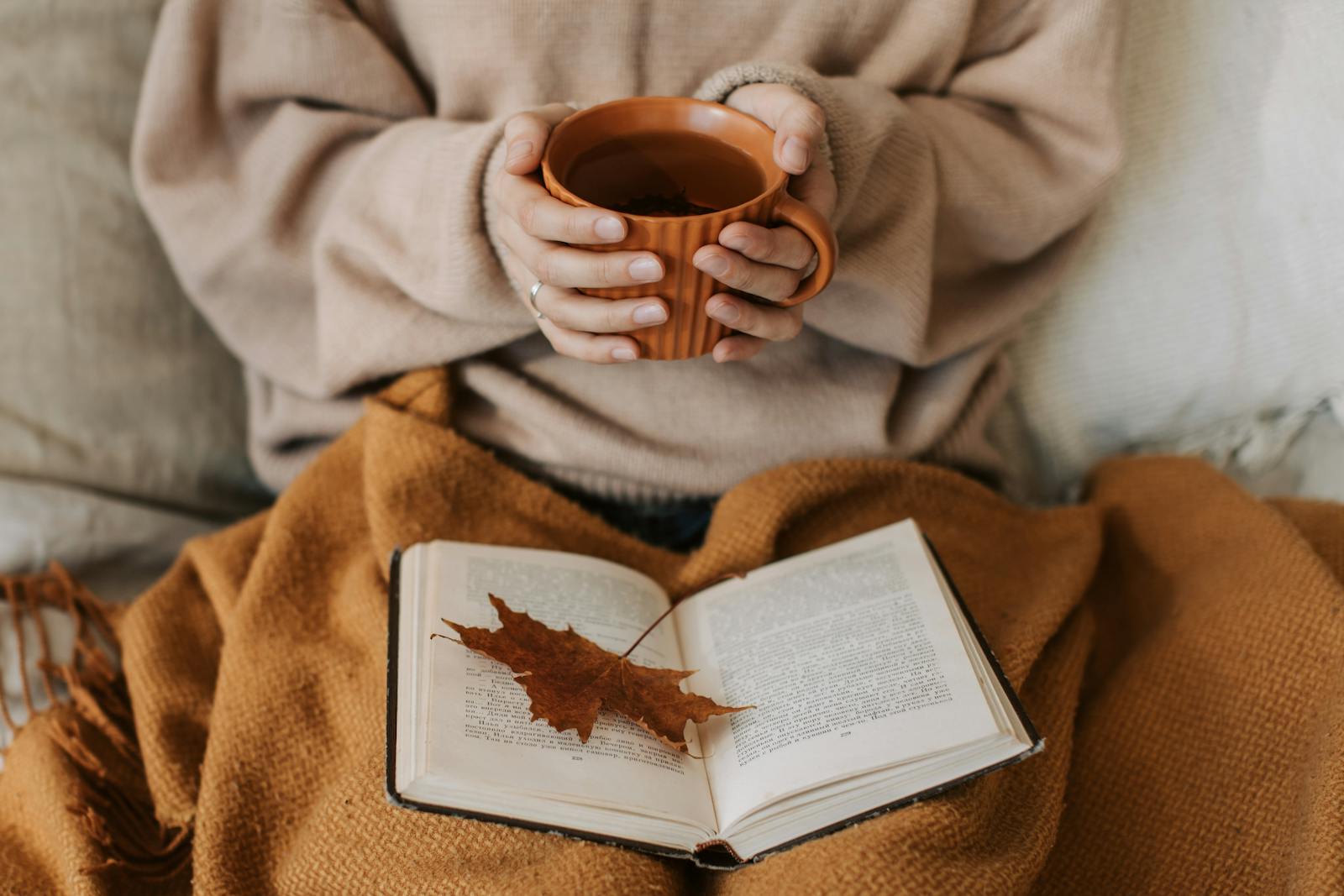 A woman enjoys a cozy autumn day, holding a tea mug over an open book with a maple leaf. skip the sales